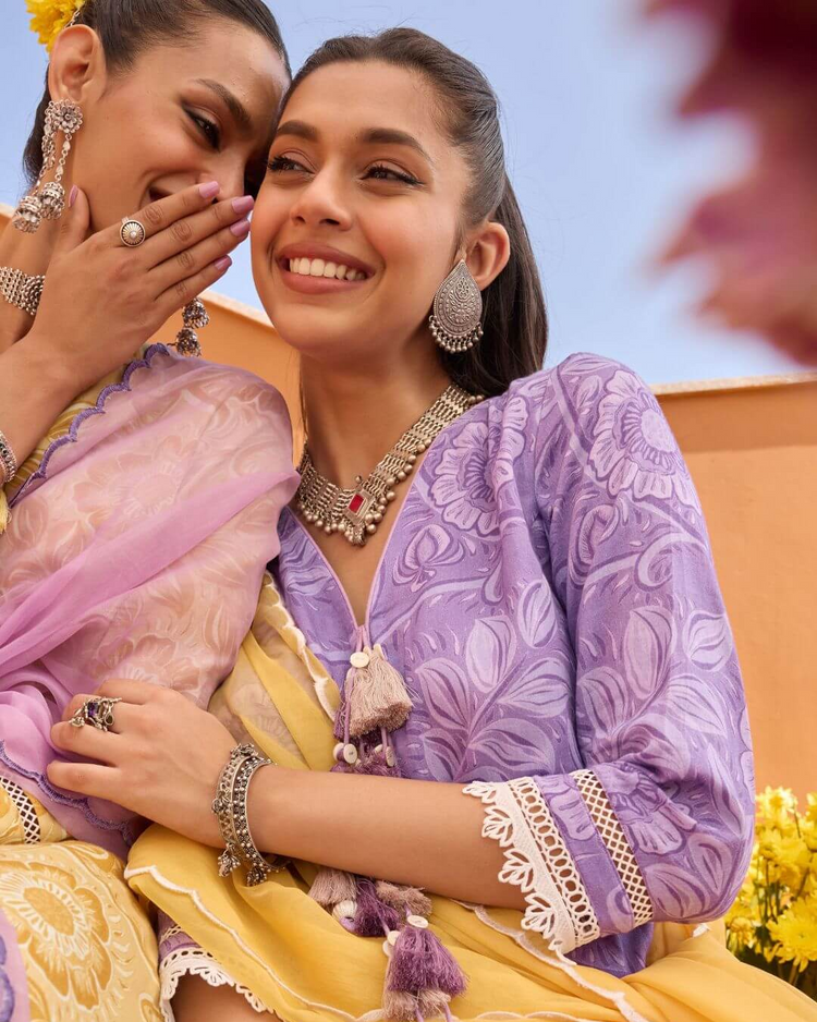Two women in traditional attire, one in purple and yellow, the other in pink and yellow, with Sadabahaar jewelry.