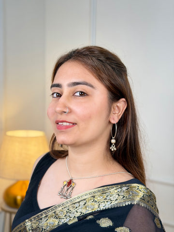 A woman wearing a black and gold embroidered traditional sari, silver necklace, and dangling earrings, photographed indoors against a light wall with a lamp in the background.