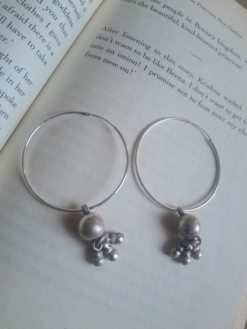 A pair of silver-tone hoop earrings with small pearl-like beads and tiny metallic charms, displayed on the pages of an open book.