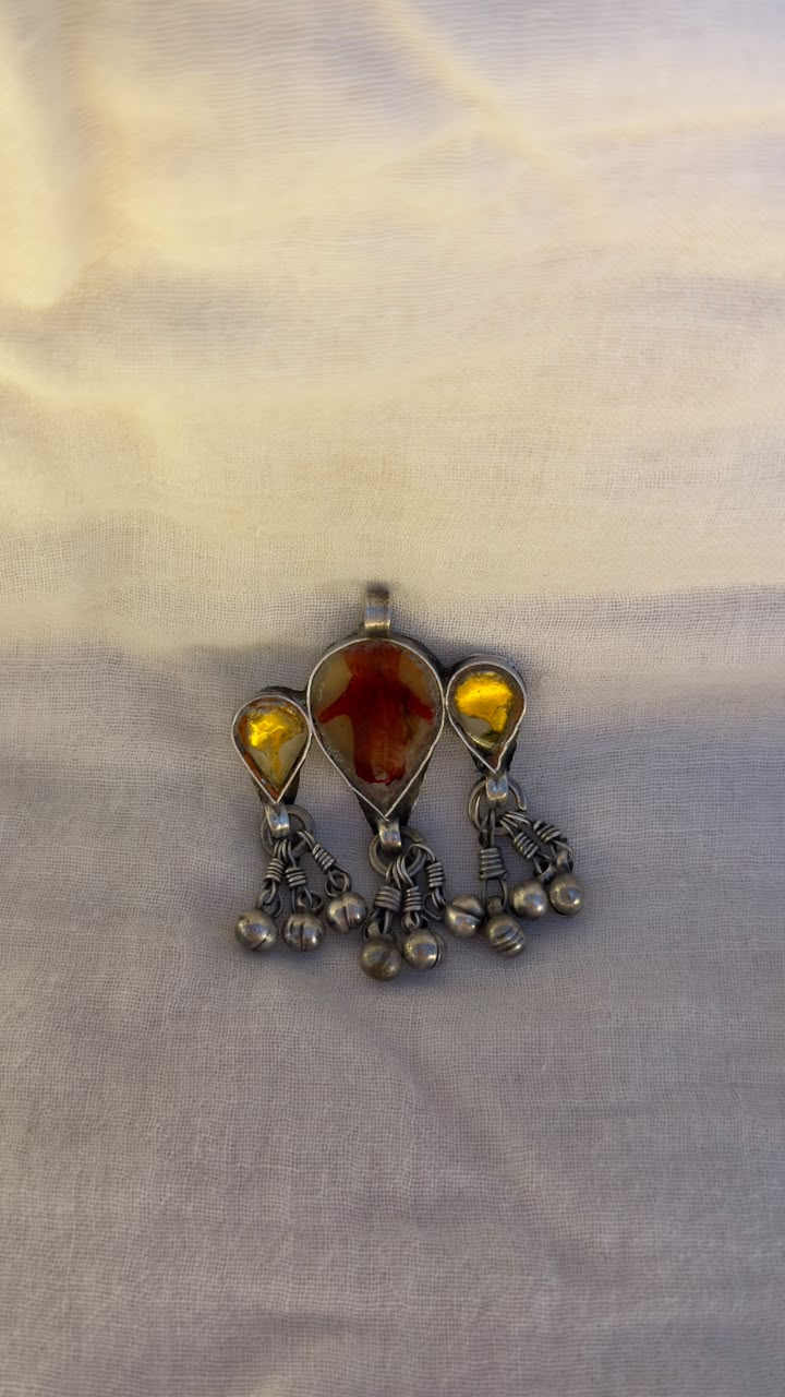 A vintage-style silver-toned pendant featuring a central teardrop-shaped amber gemstone flanked by two smaller round amber stones, with dangling silver chains and beads, displayed on a light beige fabric background.