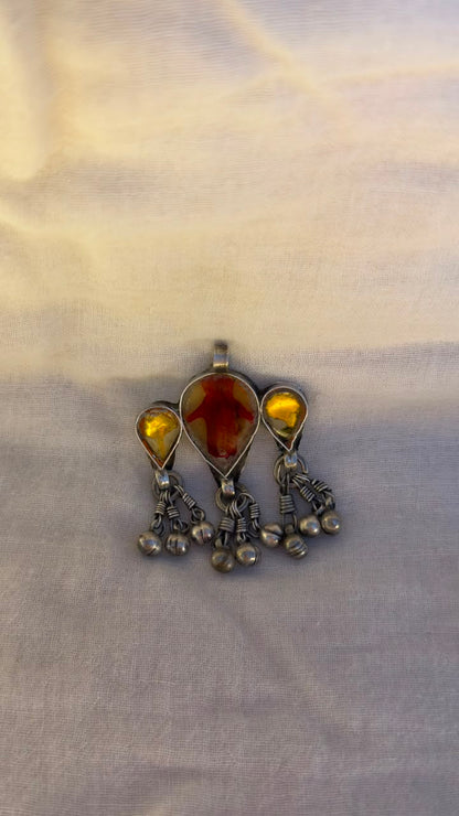 A vintage-style silver-toned pendant featuring a central teardrop-shaped amber gemstone flanked by two smaller round amber stones, with dangling silver chains and beads, displayed on a light beige fabric background.