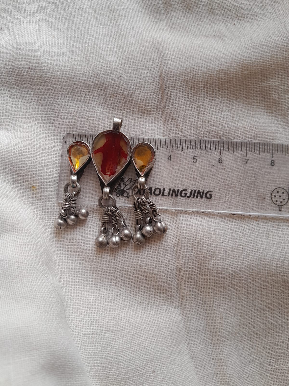 A pair of silver-toned traditional Indian pendant with teardrop-shaped amber-colored stones and dangling bead chains, shown next to a clear plastic ruler on a textured off-white fabric background.