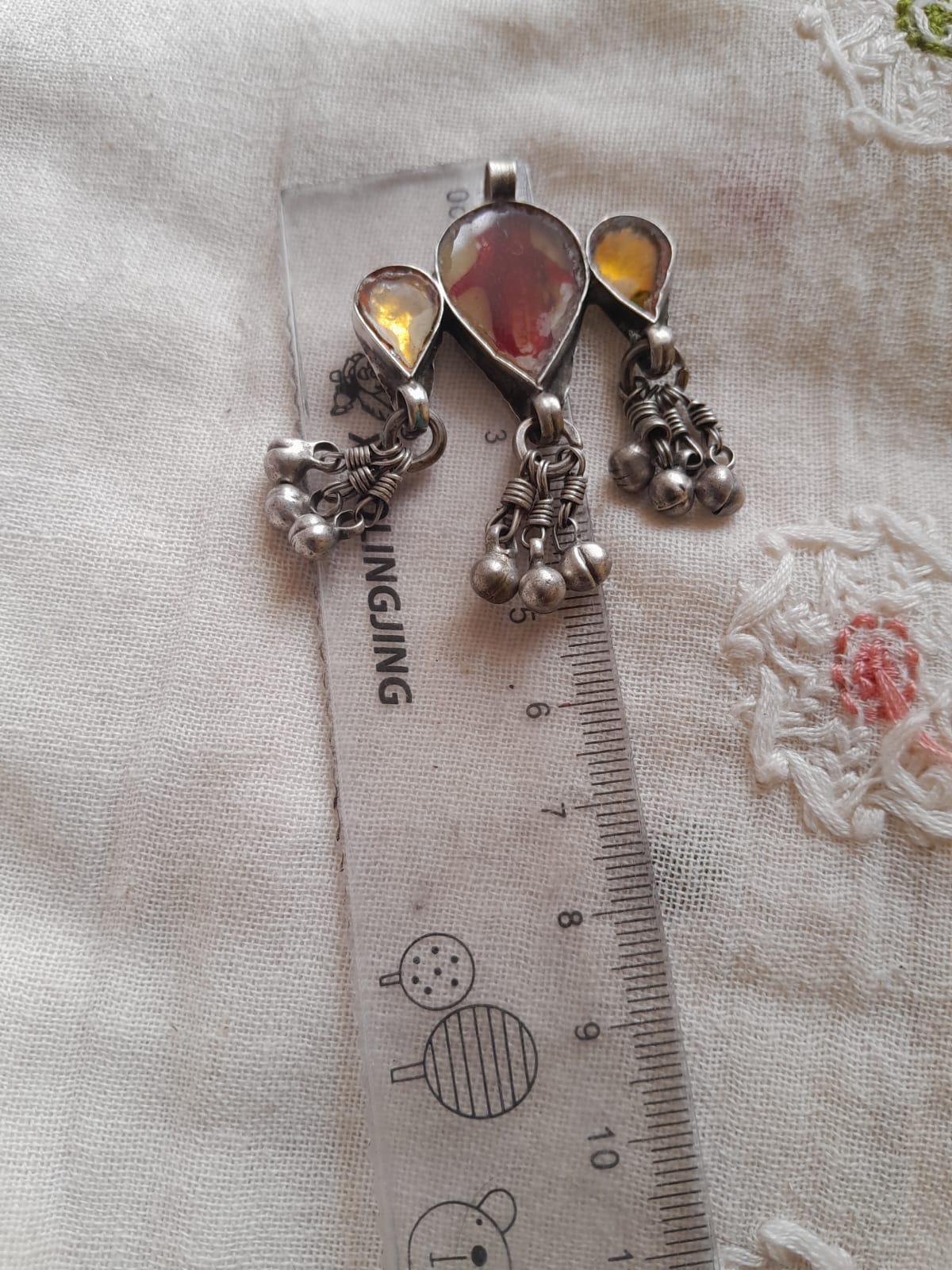 A silver-toned traditional pendant with teardrop-shaped amber stones and dangling silver bells, shown next to a ruler for scale on a textured white fabric background.