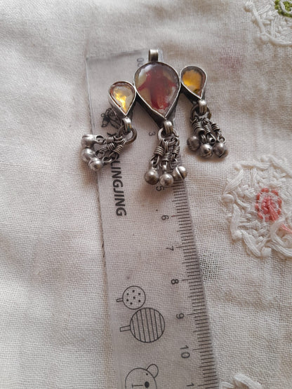 A silver-toned traditional pendant with teardrop-shaped amber stones and dangling silver bells, shown next to a ruler for scale on a textured white fabric background.