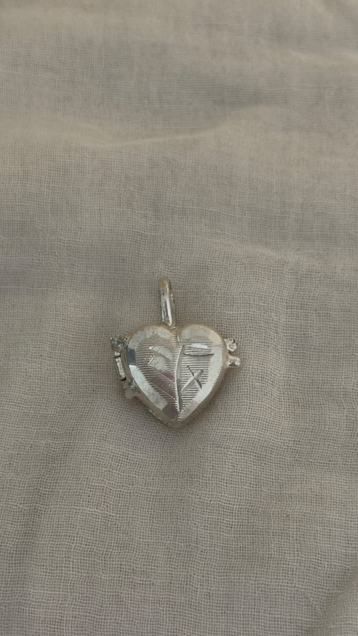 A small silver heart-shaped pendant with engraved details, resting on a textured beige fabric background.