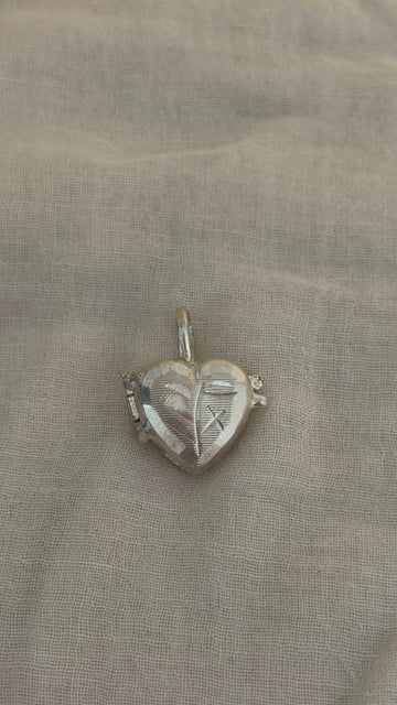 A small silver heart-shaped pendant with engraved details, resting on a textured beige fabric background.