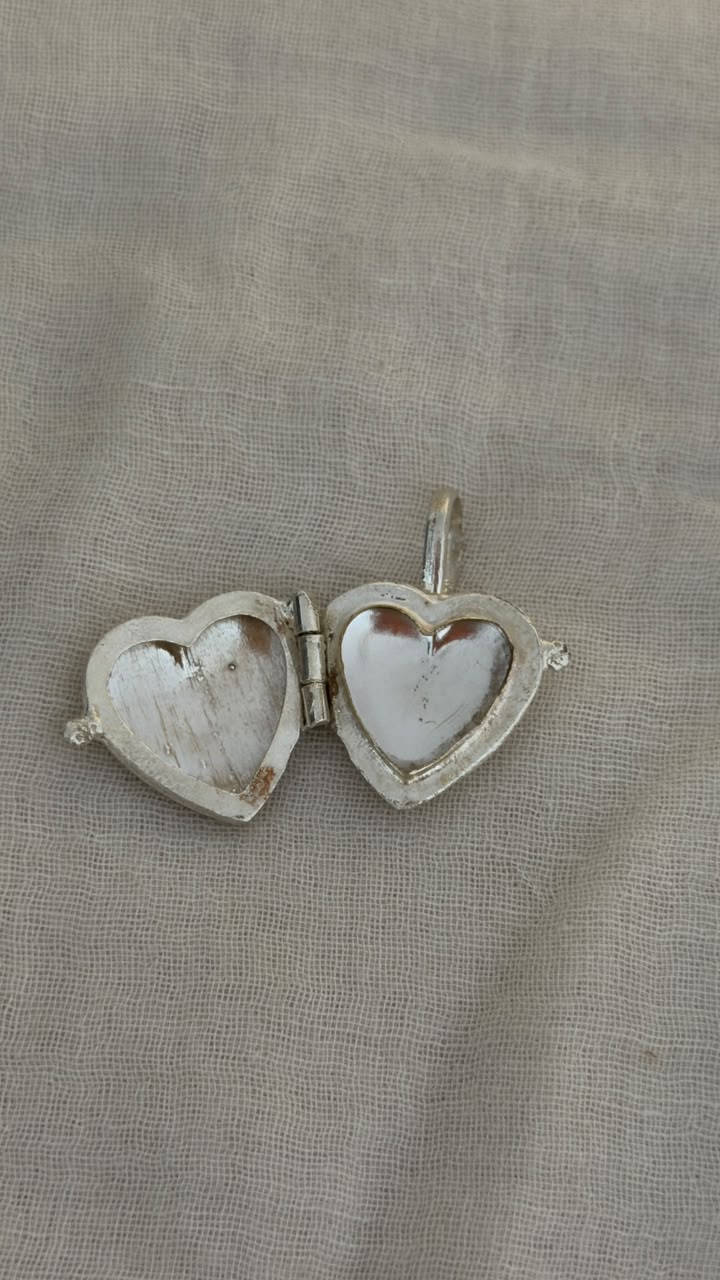 A silver heart-shaped locket pendant with a mother-of-pearl heart insert, shown open on a textured beige fabric background.