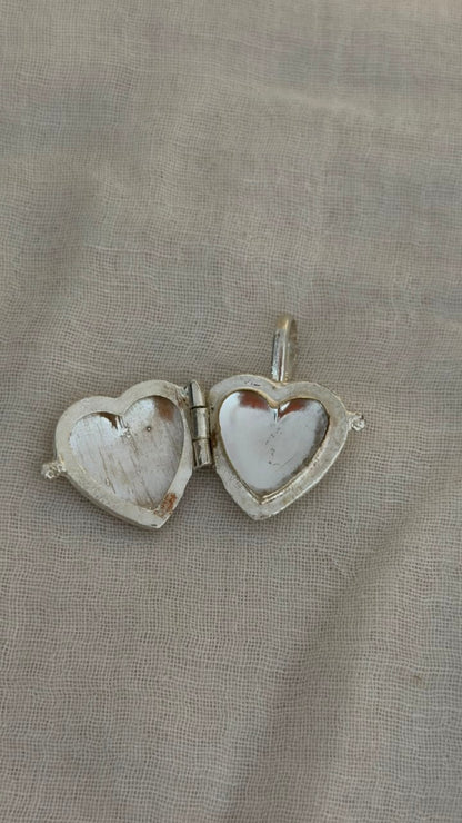 A silver heart-shaped locket pendant with a mother-of-pearl heart insert, shown open on a textured beige fabric background.