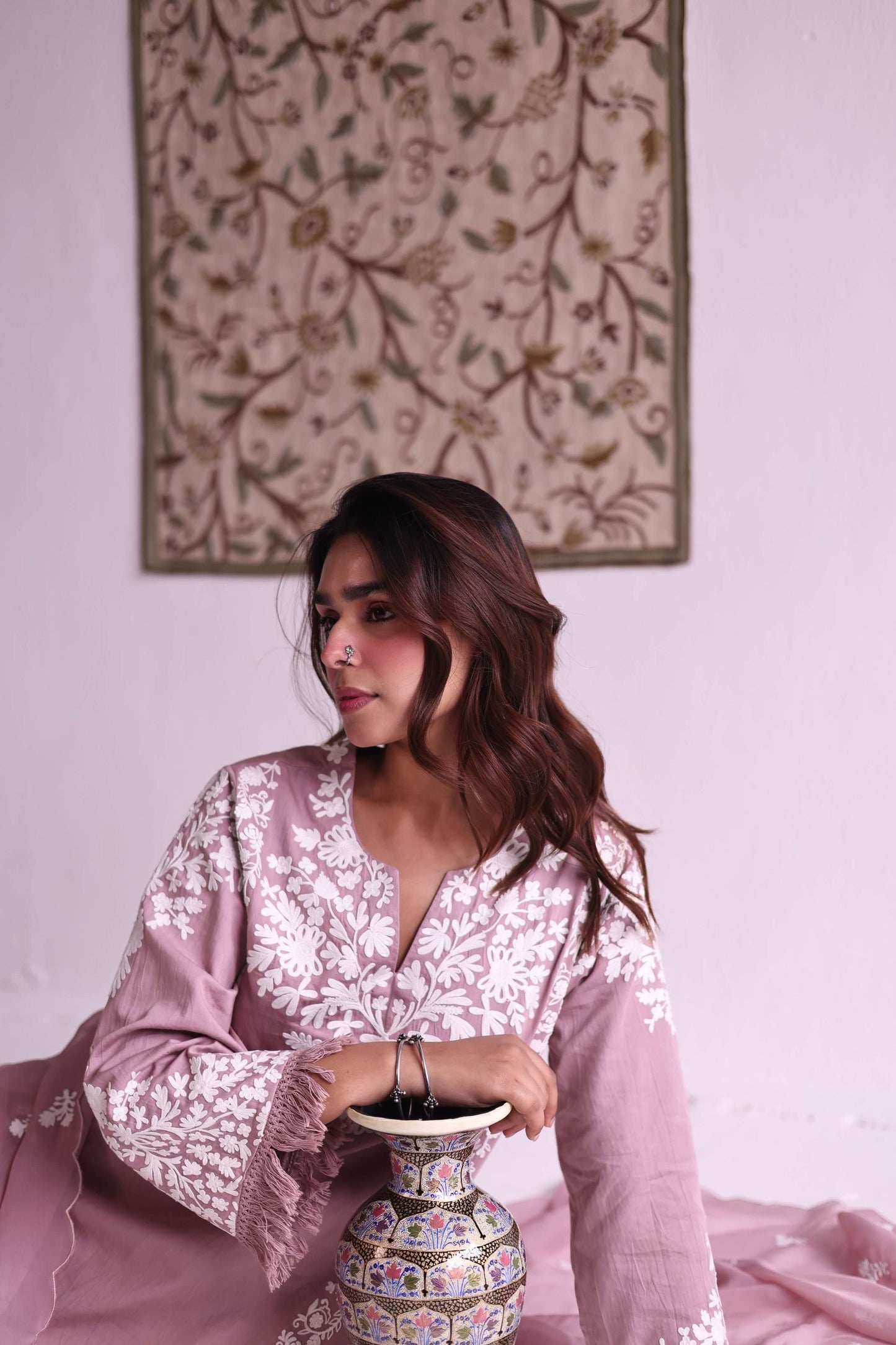 Woman in a pink embroidered outfit holding a decorative vase against a light wall with a floral painting.