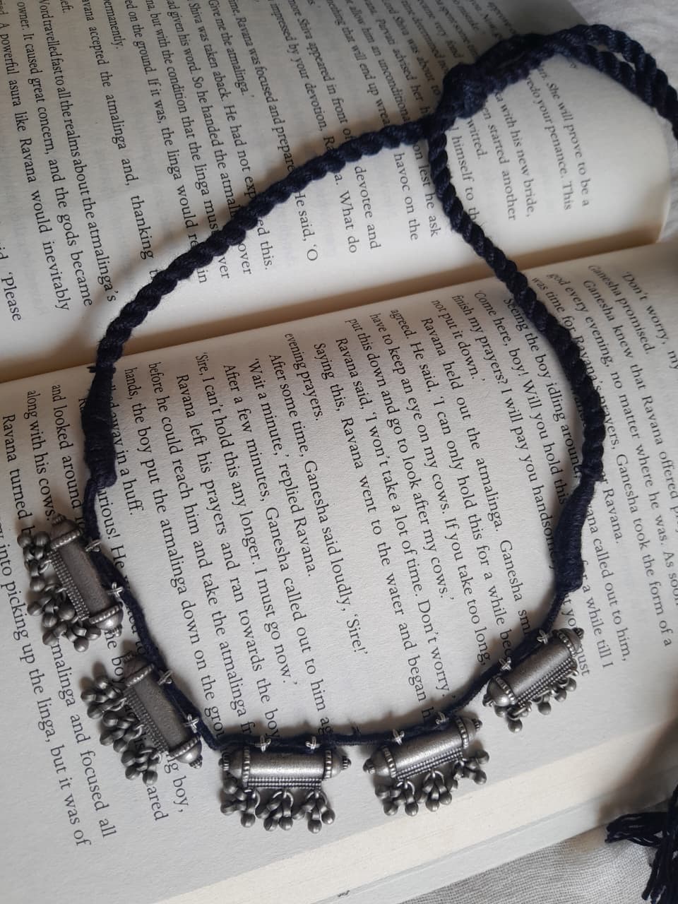 A braided cord necklace with silver-toned cylindrical and bell-shaped charms, laid flat on an open book with printed text.