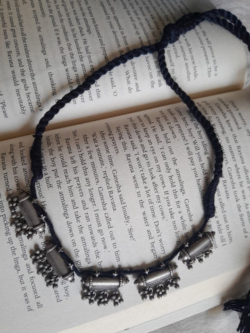 A braided cord necklace with silver-toned cylindrical and bell-shaped charms, laid flat on an open book with printed text.