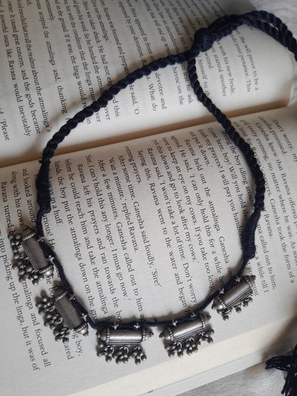 A braided cord necklace with silver-toned cylindrical and bell-shaped charms, laid flat on an open book with printed text.
