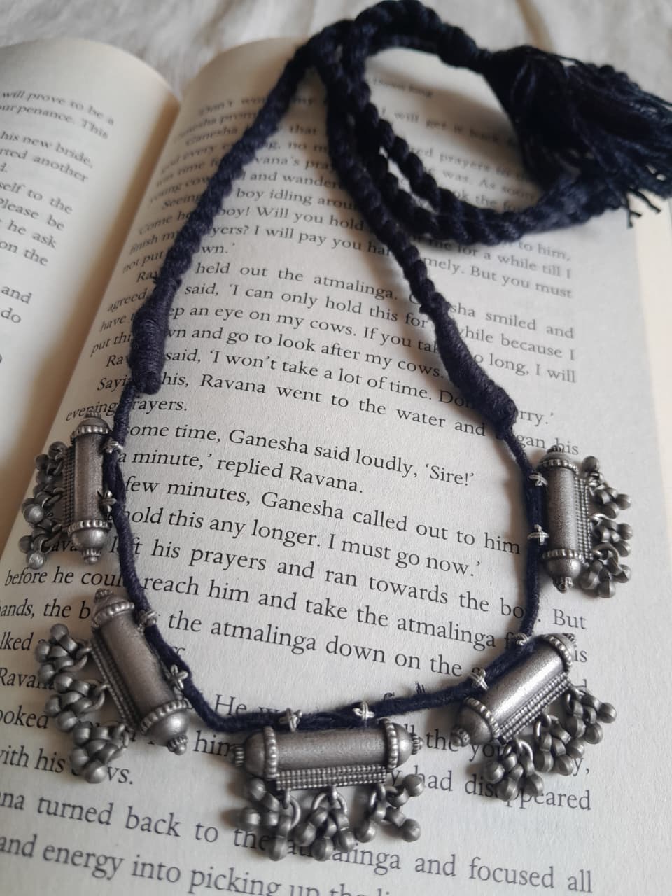 A  braided cord necklace with multiple silver-toned metal charms, including a central rectangular charm and dangling bell-like pendants, laid across an open book with printed text.