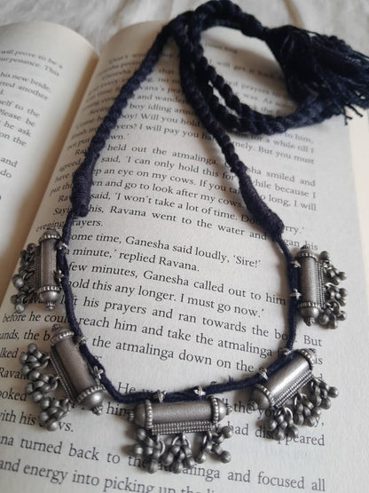 A  braided cord necklace with multiple silver-toned metal charms, including a central rectangular charm and dangling bell-like pendants, laid across an open book with printed text.