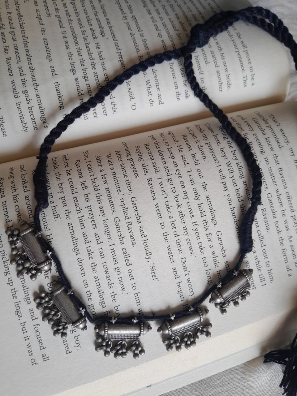 A braided cord necklace with silver-toned cylindrical and floral charms, laid out on an open book with printed text.