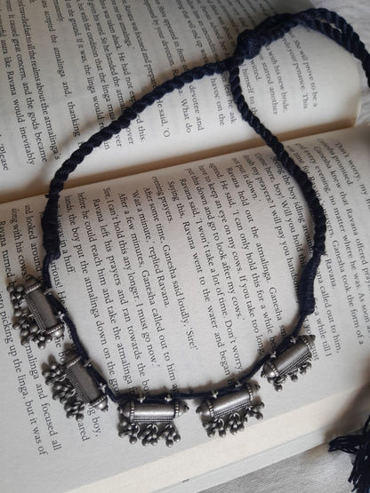 A braided cord necklace with silver-toned cylindrical and floral charms, laid out on an open book with printed text.