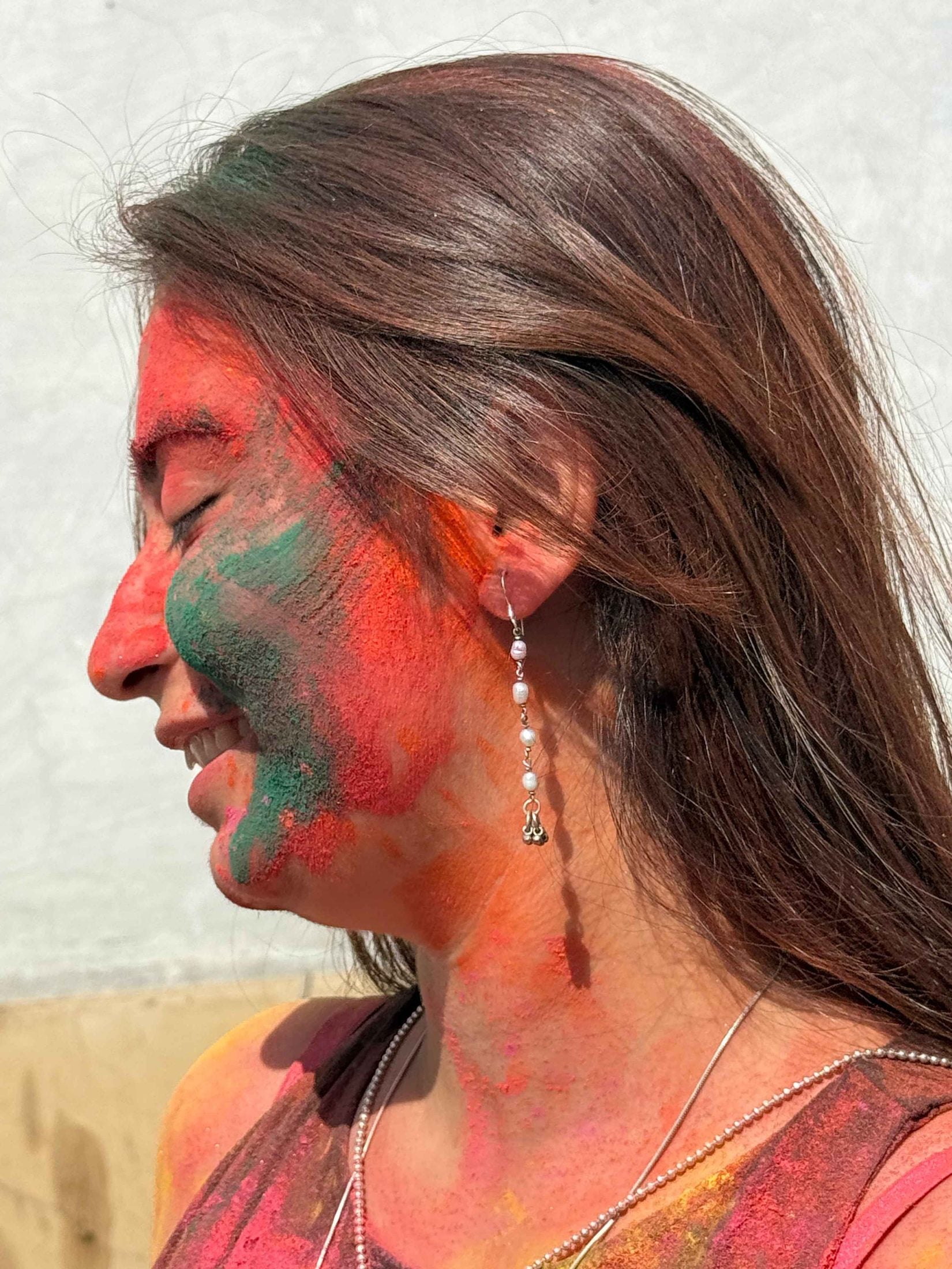 Woman with colorful face paint, likely from Holi festival, with silver pearl earrings against a neutral background.