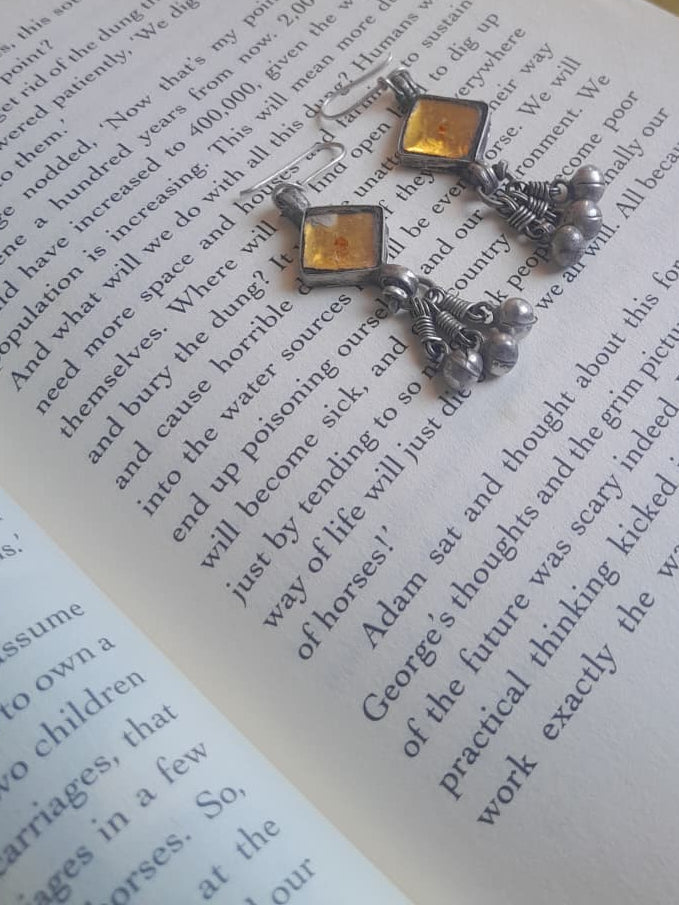 A pair of silver-toned dangling earrings with red enamel details and small bead chains, displayed on an open book over a blue patterned textile.