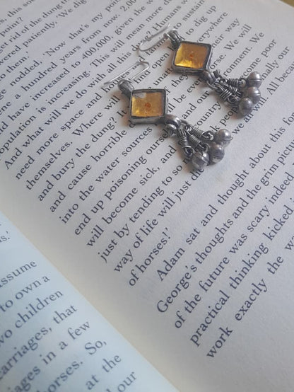 A pair of silver-toned dangling earrings with red enamel details and small bead chains, displayed on an open book over a blue patterned textile.