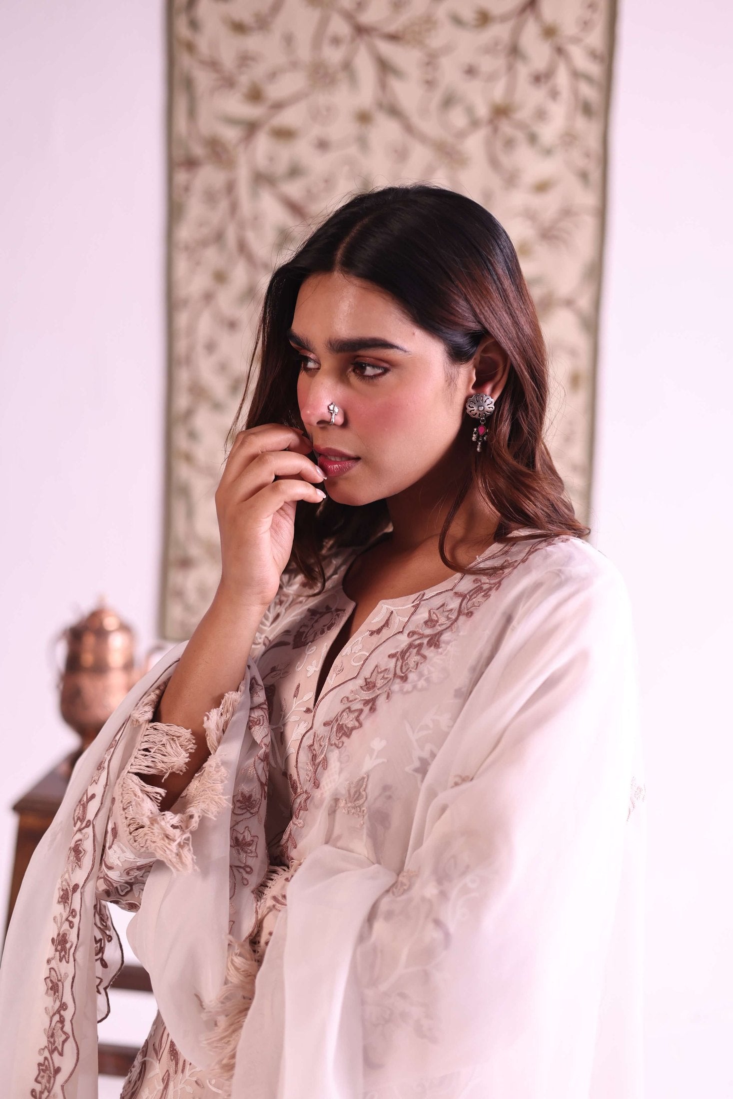 Woman in a light-colored traditional outfit with intricate patterns, standing against a decorative wall.