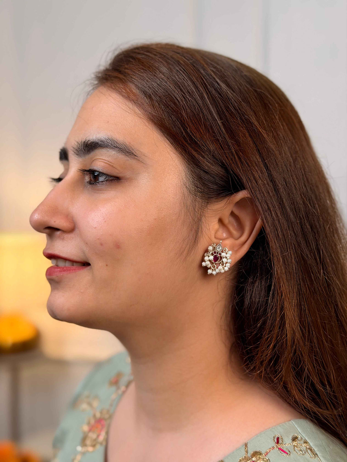 A woman shown in profile wearing a round, floral cluster stud earring with a silver-toned base, white stones, and a central deep red stone.