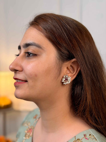 A woman shown in profile wearing a round, floral cluster stud earring with a silver-toned base, white stones, and a central deep red stone.
