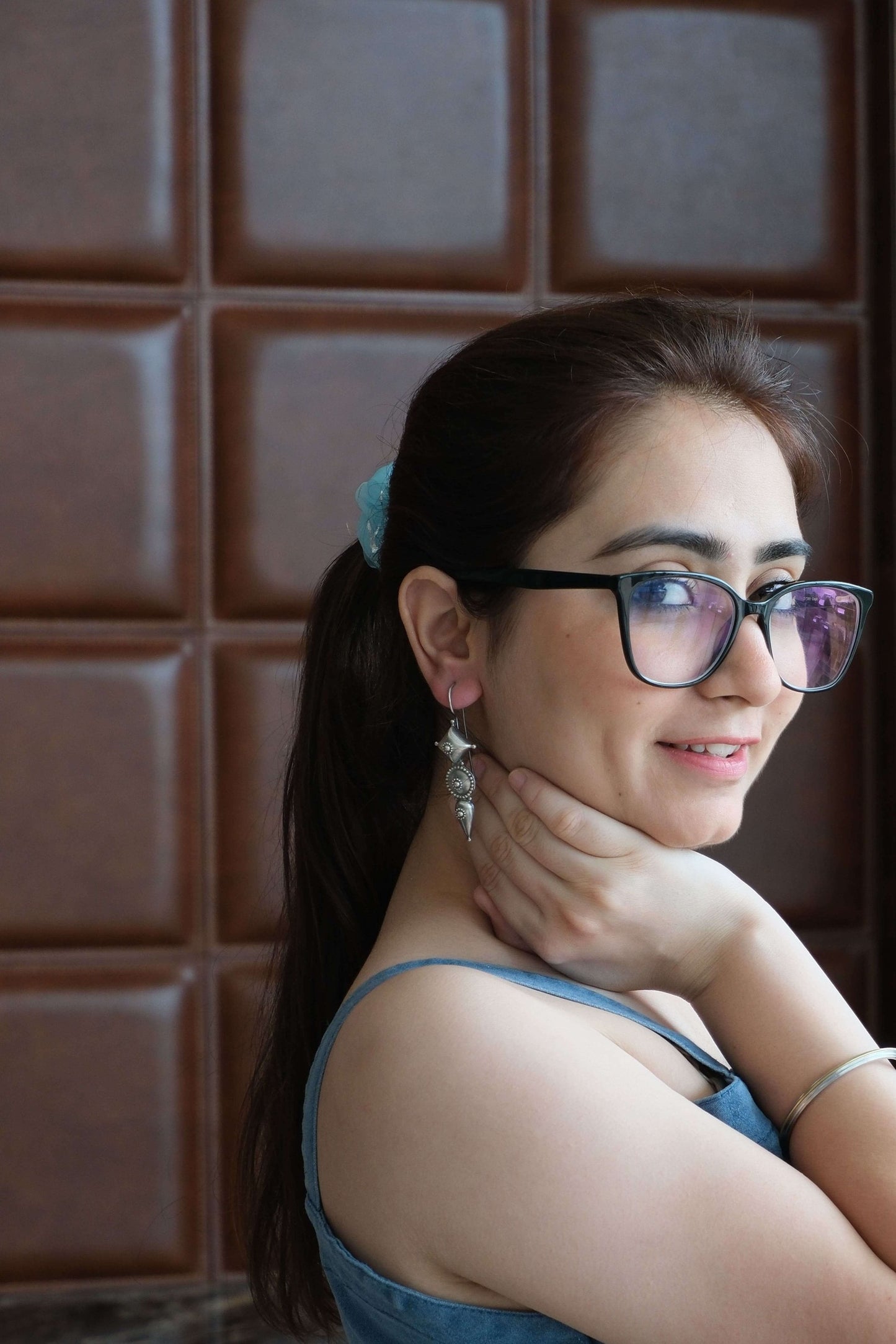 Woman wearing glasses, Gladiator Earrings, and a blue dress, sitting on a wooden floor with a brown tiled wall in the background.