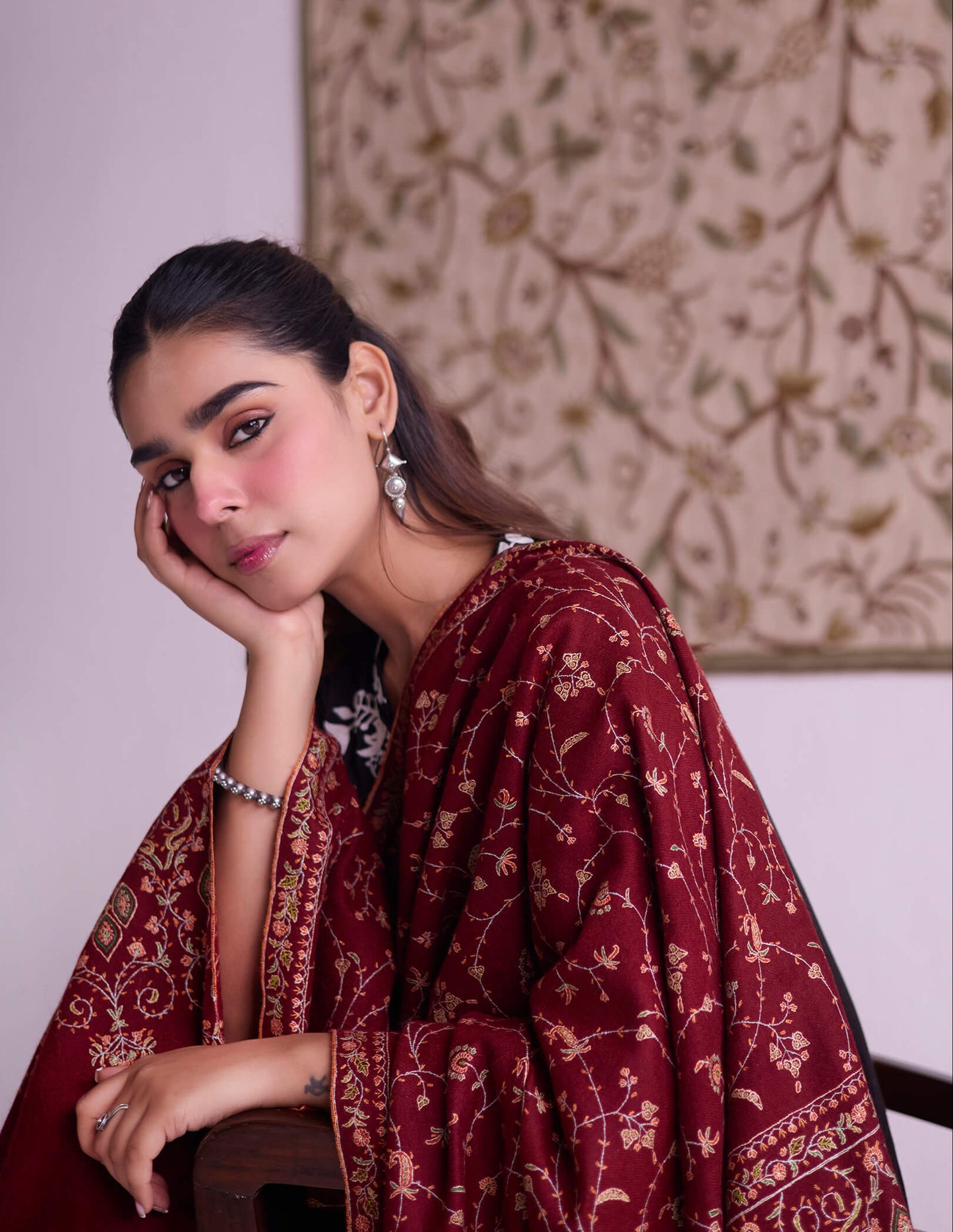 Woman wearing a maroon traditional outfit with intricate patterns, sitting against a neutral background.
