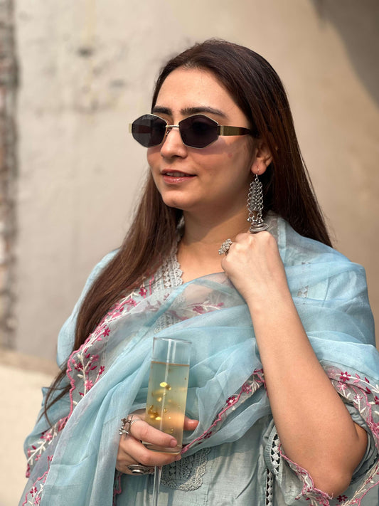 Woman in a light blue saree with floral patterns, holding a glass, against a neutral background.