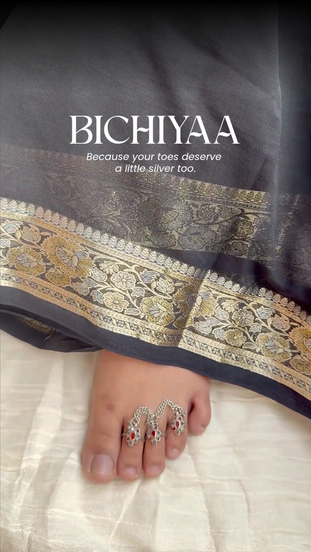 A close-up of a foot wearing ornate silver toe rings with red and white stone accents, under draped black fabric with a gold patterned border. The text 'BICHIYAA' is printed on the fabric.