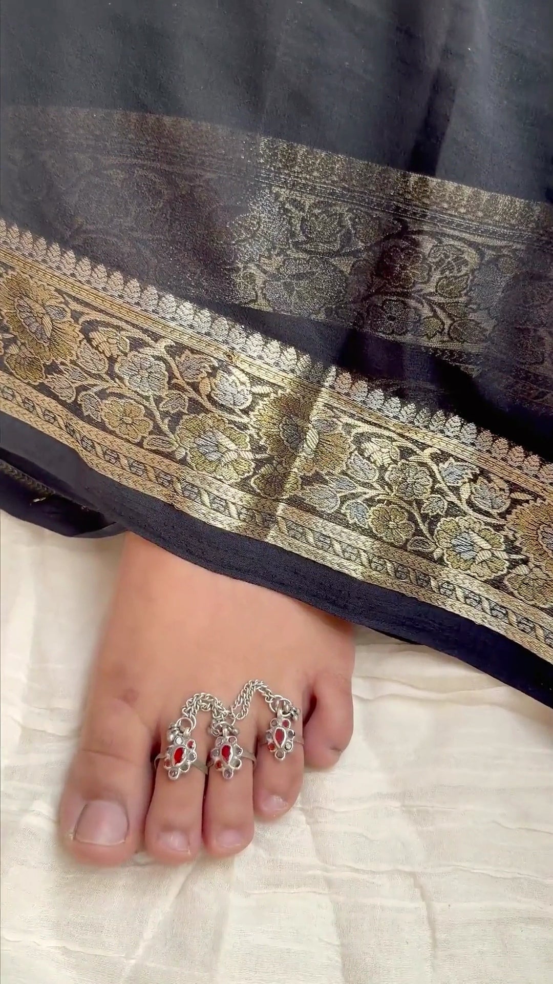 A close-up of a foot wearing three silver toe rings with red stone accents, positioned under the hem of a black and gold patterned saree on a light beige fabric surface.