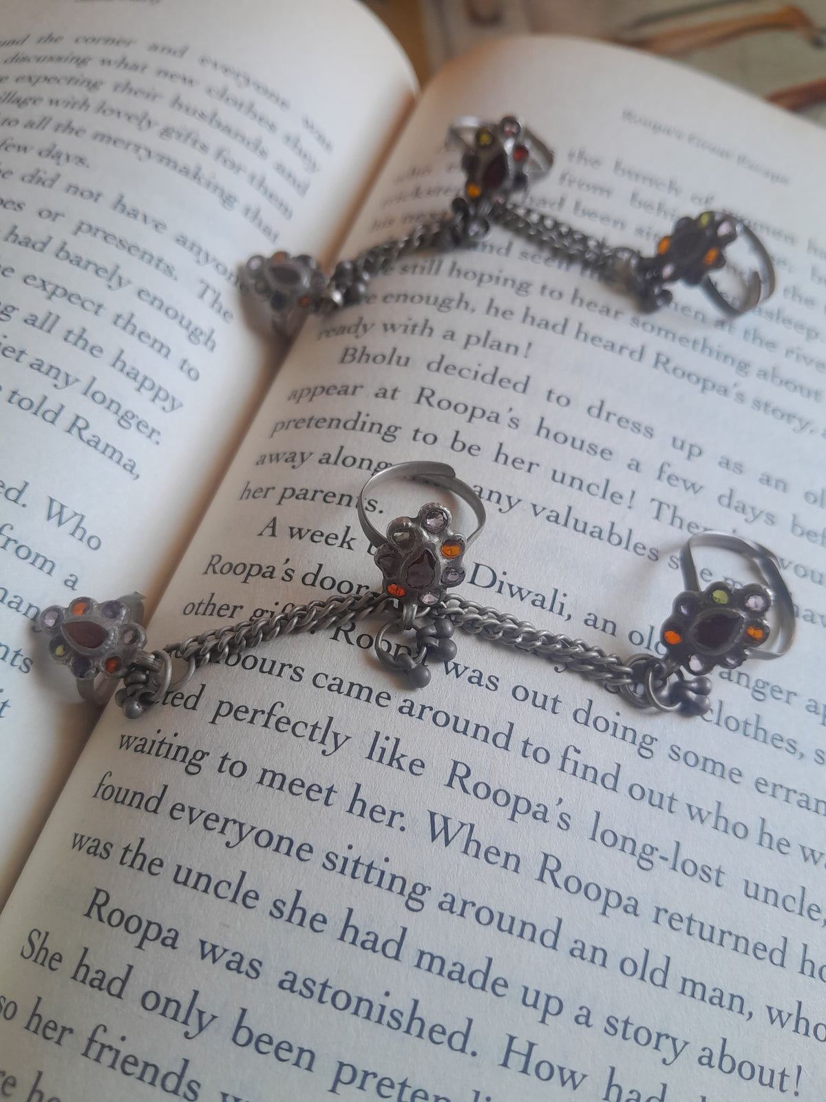 A close-up of a silver-toned chain necklace with a detailed pendant, laid across the pages of an open book. The pendant features a dark, ornate design with small orange and red gemstone accents.