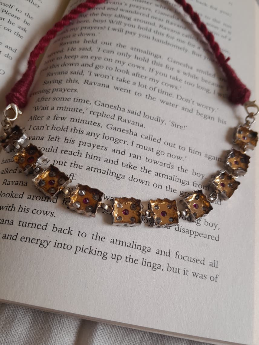 A close-up of a necklace with gold-toned floral beads and red gemstone accents, laid across an open book with printed text. A red braided cord bracelet is partially visible underneath.