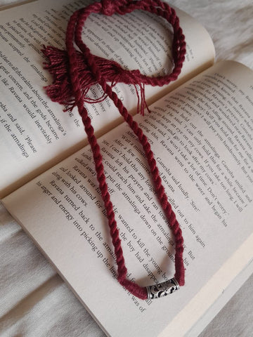 A dark red braided cord bracelet with tassel ends and a silver-toned bead, laid over the pages of an open book on a light fabric surface.