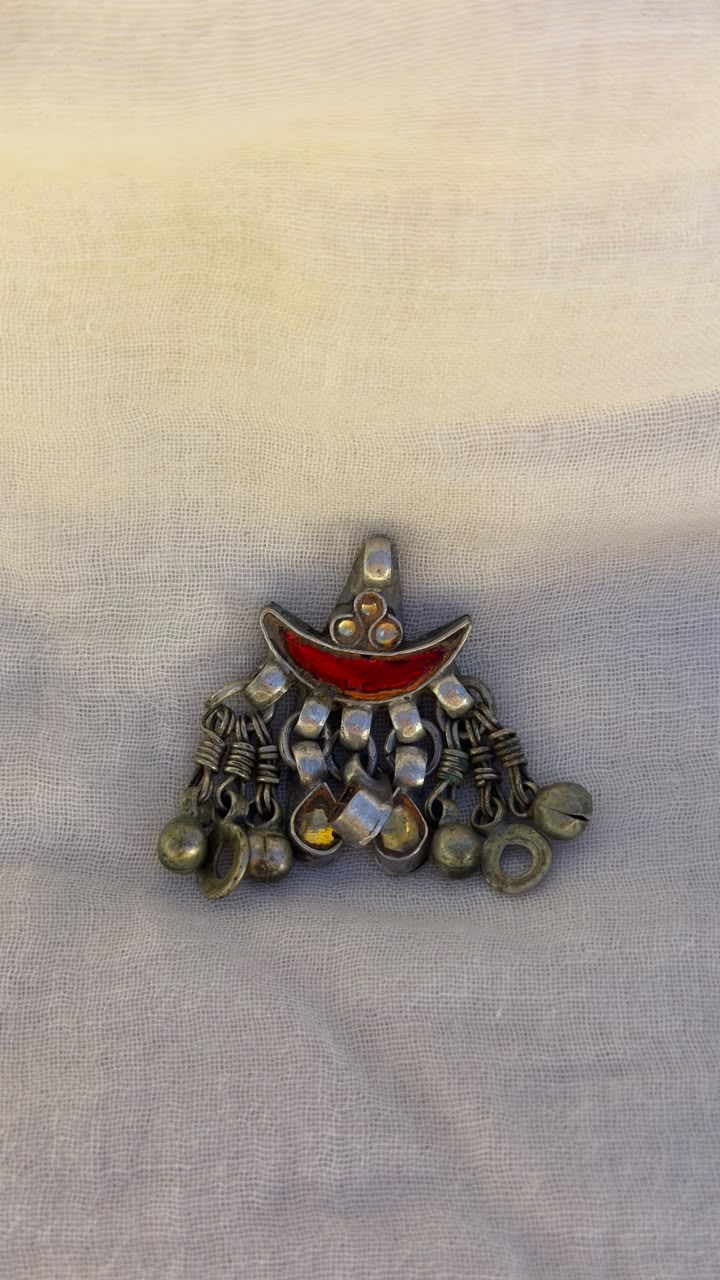 A vintage-style silver-toned crescent moon pendant brooch with a red enamel center and dangling silver bells, displayed on a light beige textured fabric background.