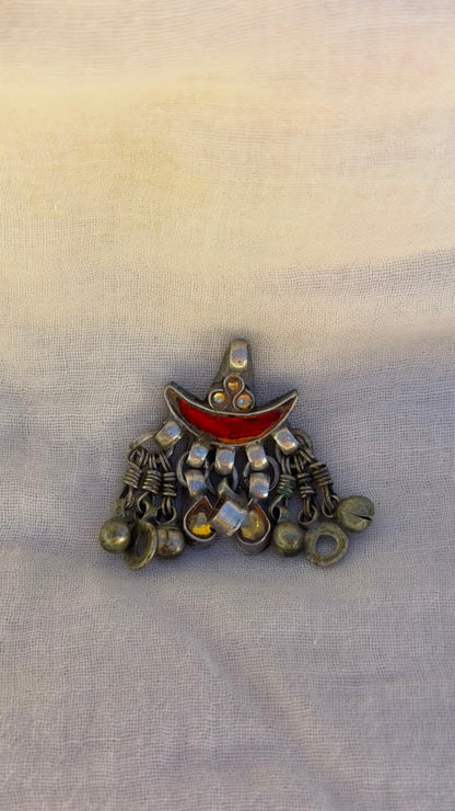 A vintage-style silver-toned crescent moon pendant brooch with a red enamel center and dangling silver bells, displayed on a light beige textured fabric background.