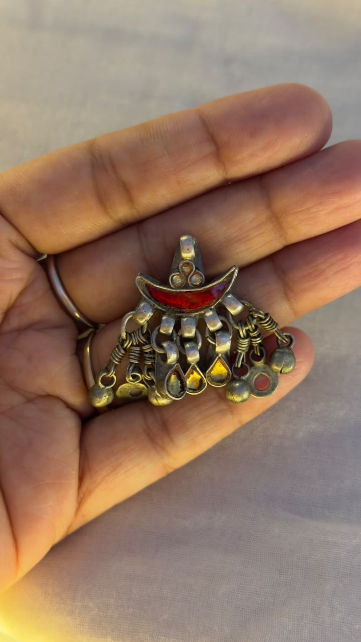 A hand holding a small ornate pendant with a gold-toned crescent moon and dangling bead-like details, featuring a red inlay at the top, against a soft gray background.