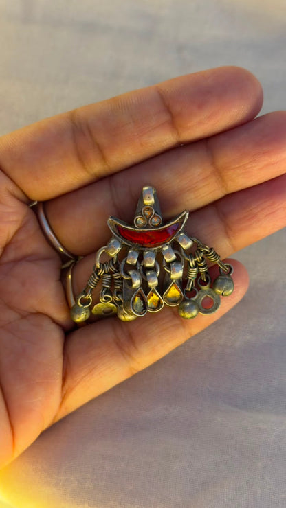 A hand holding a small ornate pendant with a gold-toned crescent moon and dangling bead-like details, featuring a red inlay at the top, against a soft gray background.
