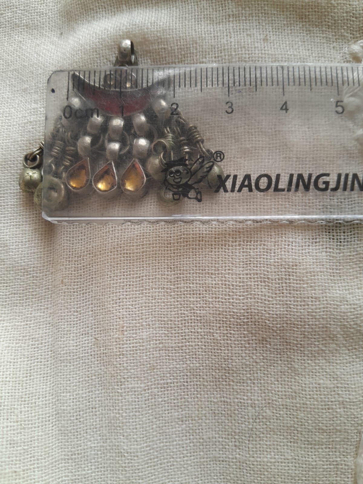 A close-up of a silver-toned beaded jewelry piece with teardrop charms resting on a clear plastic ruler over a beige fabric background.
