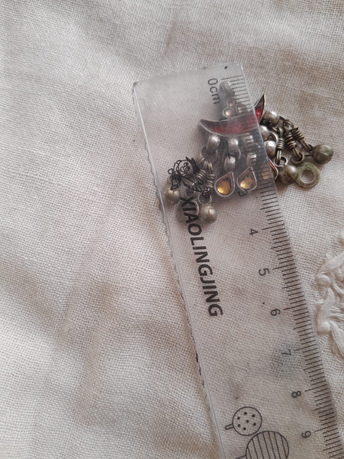 A small cluster of metallic jewelry pendant, including silver-toned beads, chain links, and a small red-and-silver charm, placed next to a clear plastic ruler on a light fabric background.