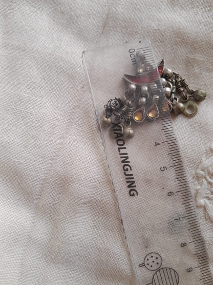 A small cluster of metallic jewelry pendant, including silver-toned beads, chain links, and a small red-and-silver charm, placed next to a clear plastic ruler on a light fabric background.