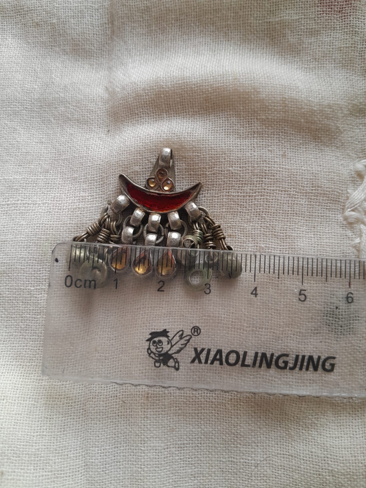 A small silver-toned crescent moon pendant with dangling bead-like details and a red center, placed above a clear ruler on a textured off-white fabric background.