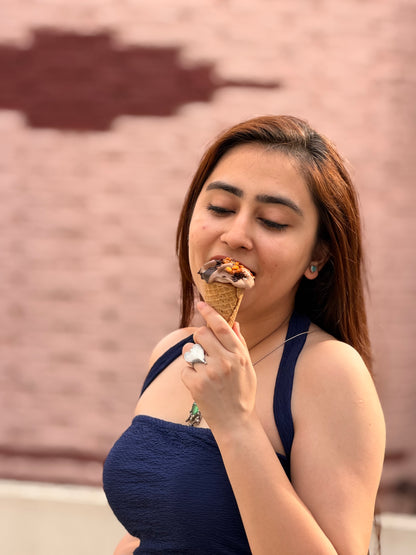 A woman with long brown hair wearing a dark blue halter top is eating a chocolate ice cream cone in front of a pinkish brick wall.