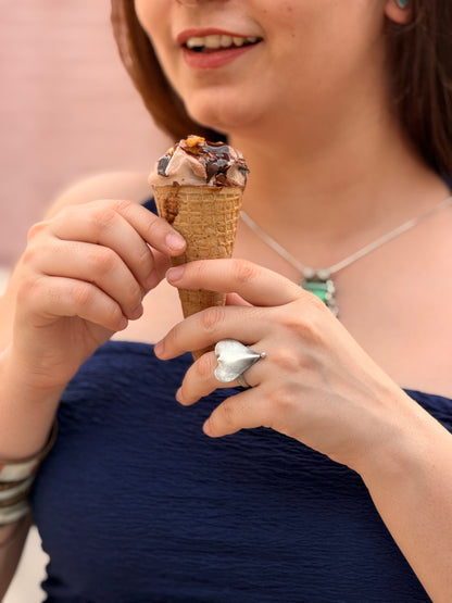 A close-up lifestyle shot of a woman holding a chocolate ice cream cone, wearing a textured navy blue top, a silver necklace, and a large silver ring.