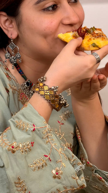 A close-up lifestyle shot of a woman wearing a sage green embroidered kurta and silver jewelry, eating a piece of pizza with her hand.