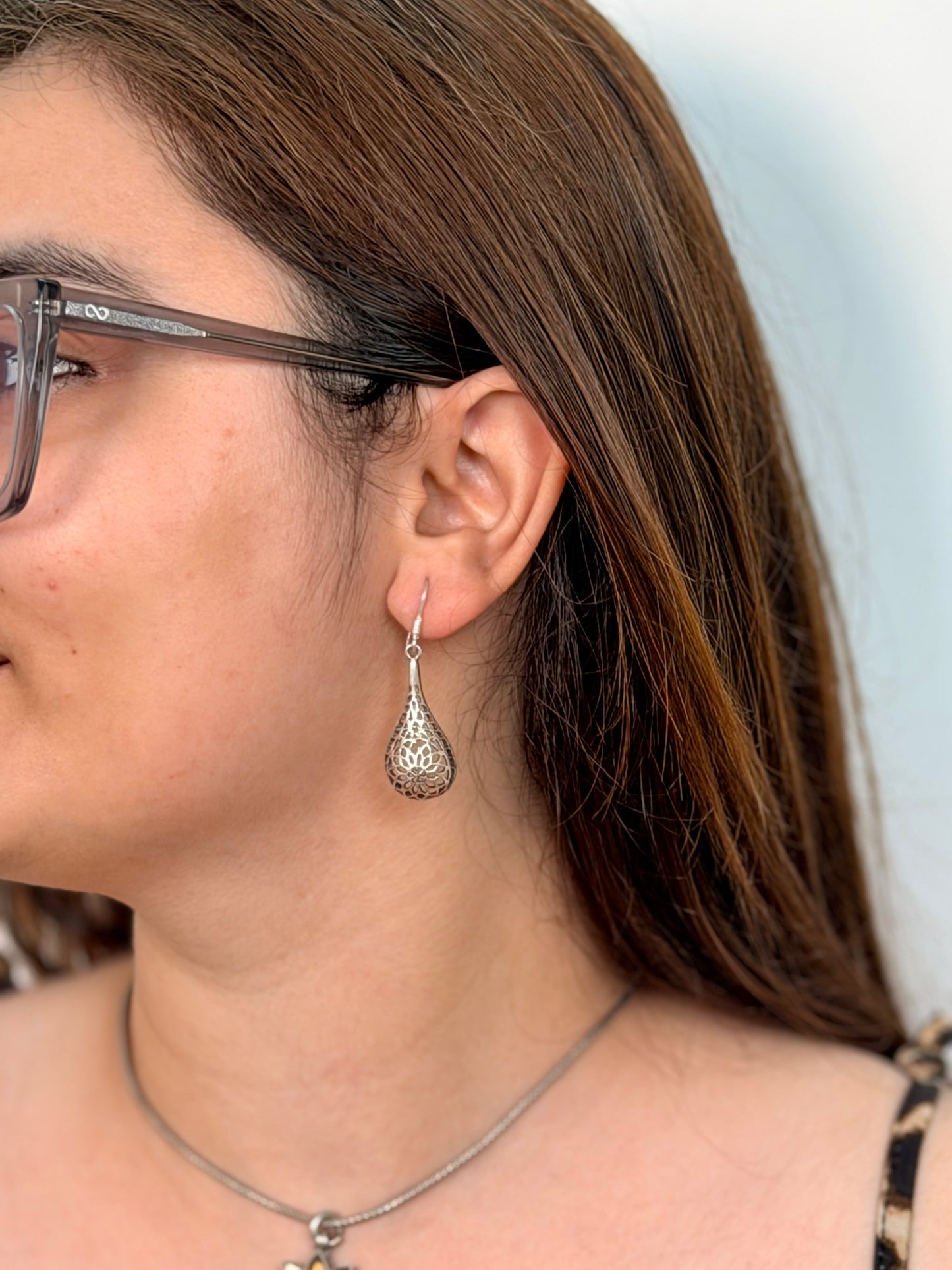 A close-up side profile of a woman wearing a silver teardrop dangle earring with intricate filigree detailing, along with a silver chain necklace and glasses.