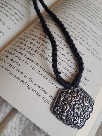 A dark braided cord necklace with a square, heavily engraved silver-toned pendant featuring a floral and bird motif, laid across the pages of an open book.