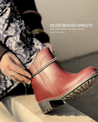 Red boot with silver beaded anklets worn by a person sitting on a stone surface.