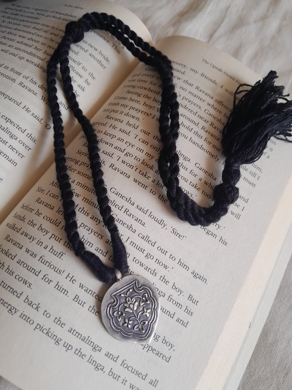 A black braided cord necklace with a silver-toned shield pendant and black tassel, laid over an open book page.