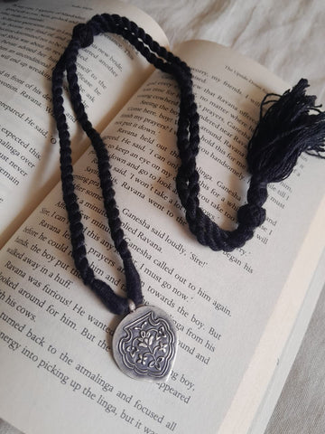 A black braided cord necklace with a silver-toned shield pendant and black tassel, laid over an open book page.